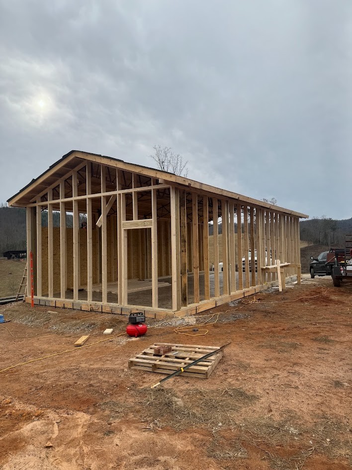 Wood-framed outbuilding with wall studs, roof trusses, and partial sheathing on a hillside