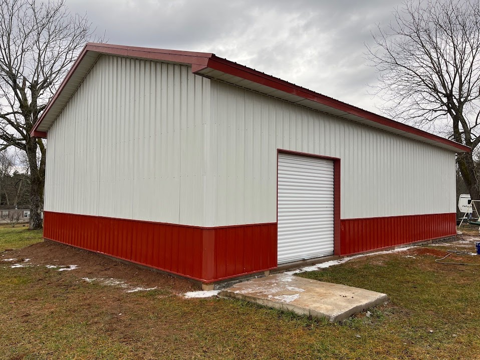 White corrugated metal utility building with deep red wainscoting, a red gable roof, and a single white roll-up door on a concrete landing pad
