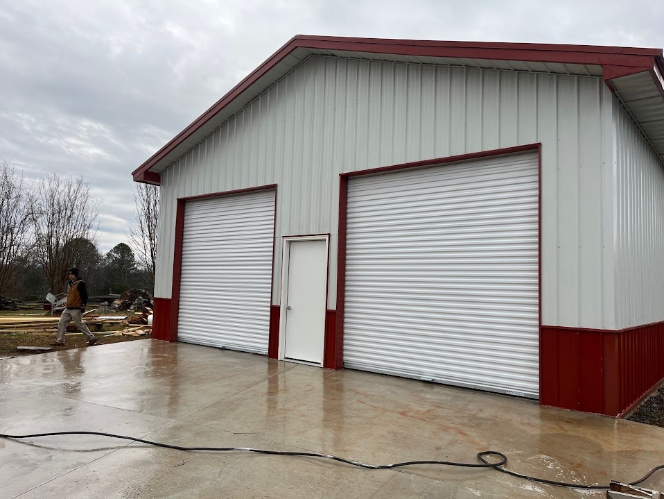 Two-bay metal garage with white vertical siding, red wainscoting and roof trim, two white roll-up doors, and a centered entry door on a concrete apron