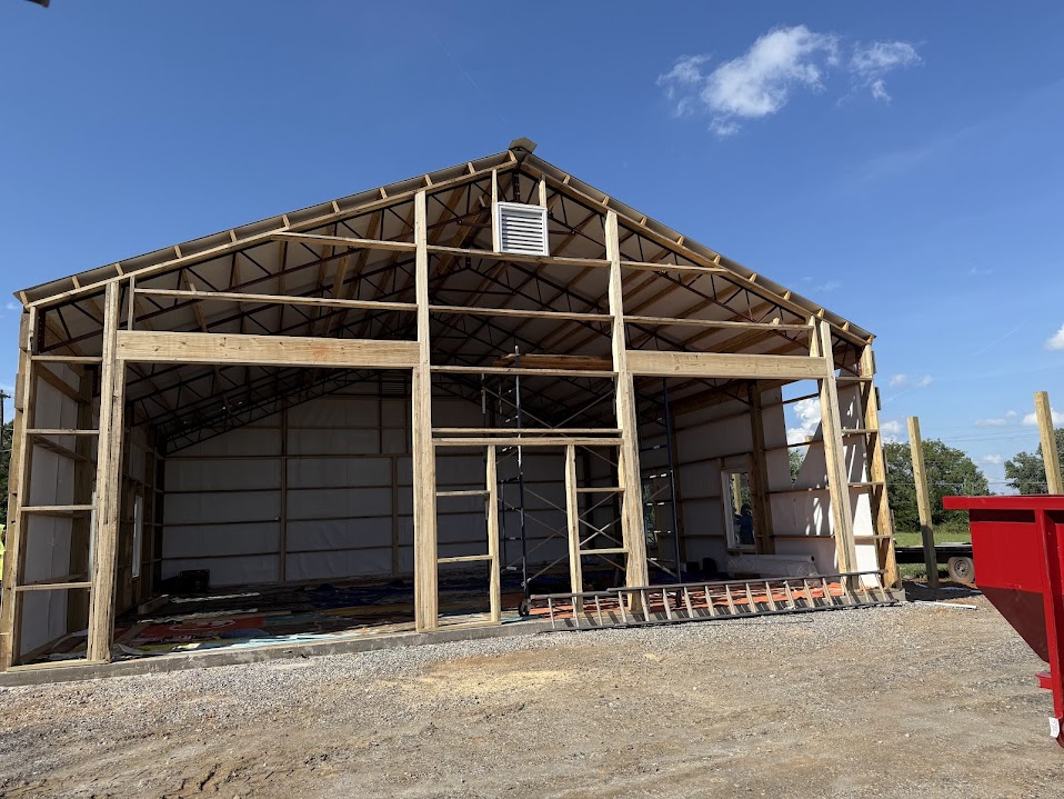 Pole barn under construction with exposed lumber framing, roof trusses, white vapor barrier insulation, and open rough door frames