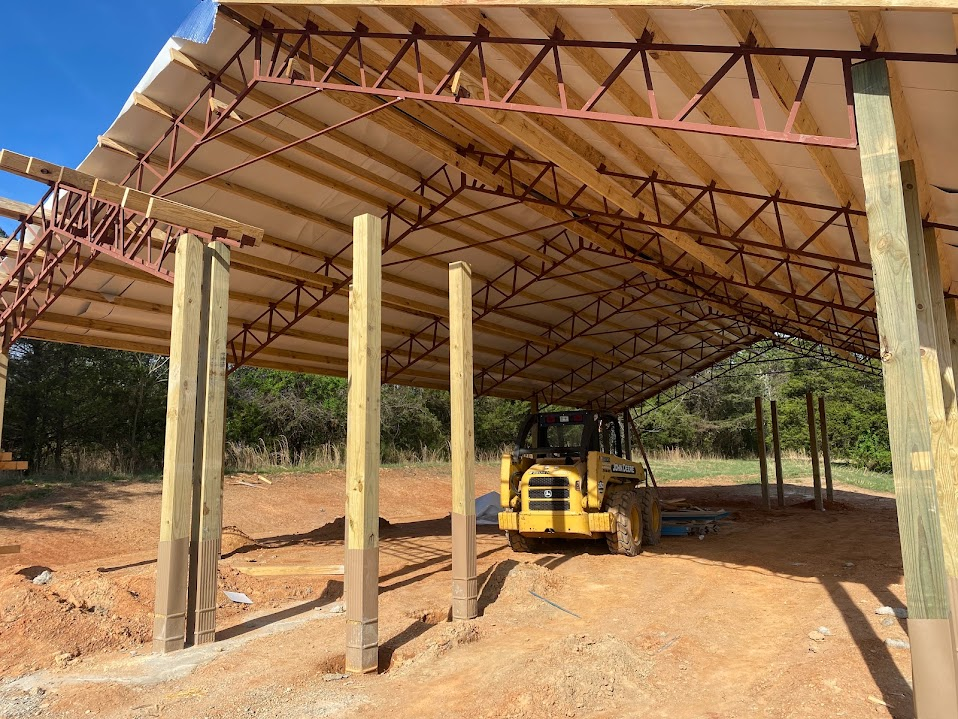 Interior of a post-frame pole barn with timber posts, steel open-web trusses, wood purlins, and a skid steer loader