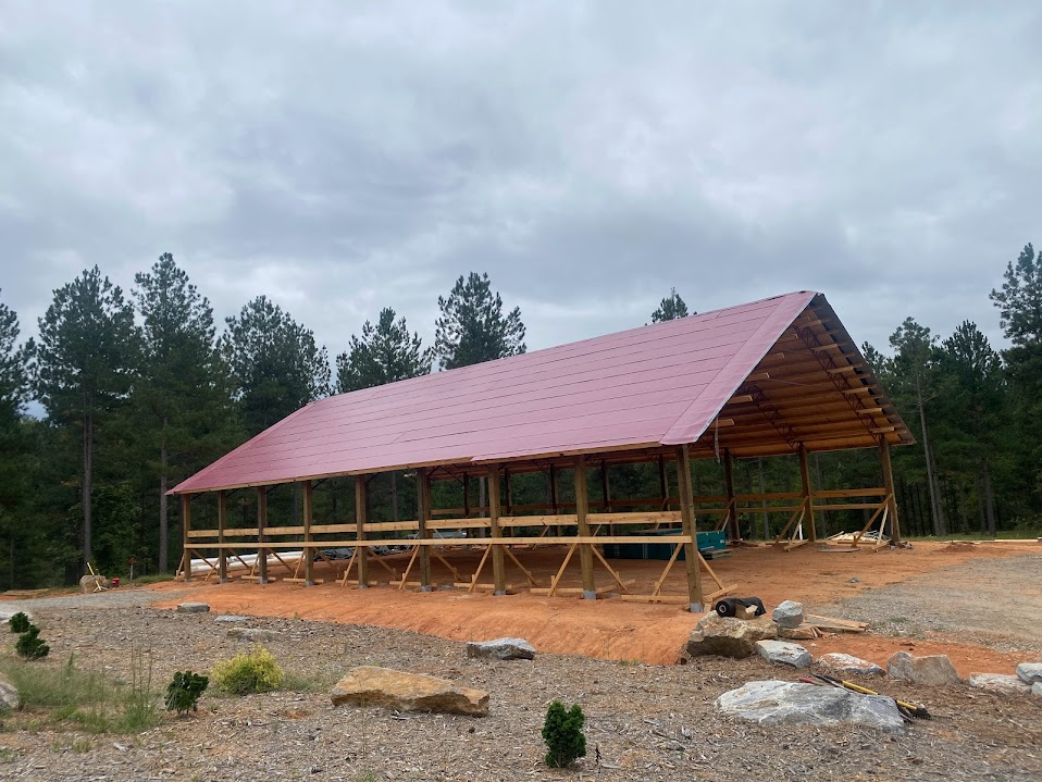 Open post-frame pole barn with timber posts and a burgundy metal roof on a cleared clay building pad