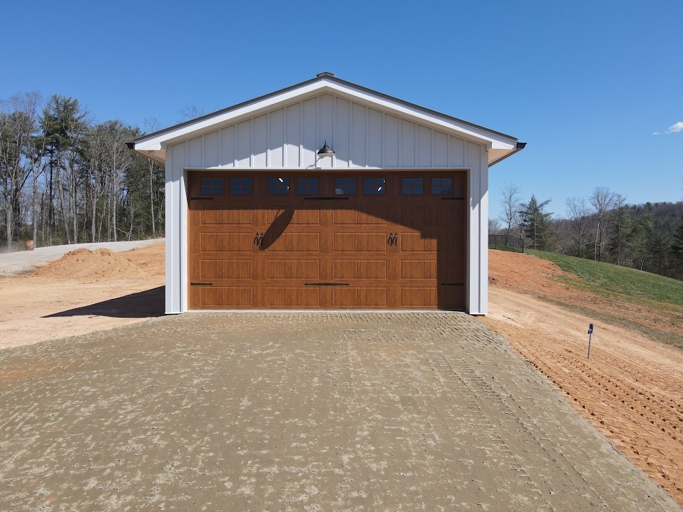Garage with white board-and-batten siding and a dark metal roof with a two-car wood-grain carriage-style door on a graded rural driveway
