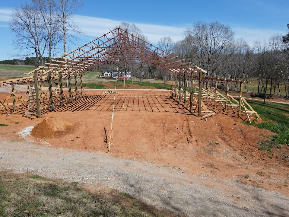 Pole barn frame with treated wood posts and red-oxide steel roof trusses on a graded clay site